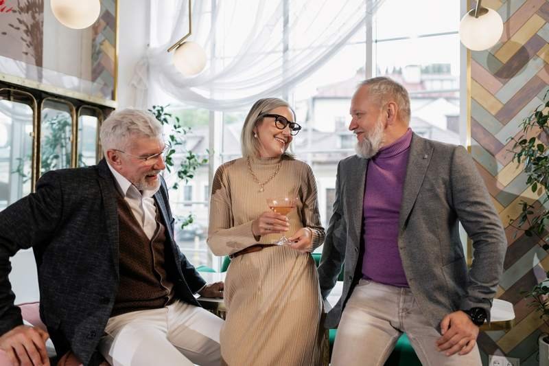 Trois amis aux cheveux blancs, élégamment habillés, trinquent et rient ensemble dans un café lumineux.