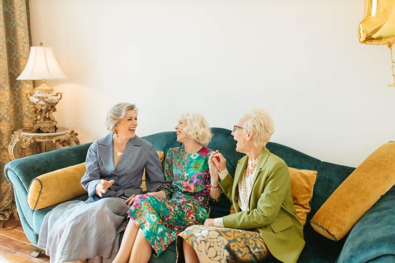 Trois femmes âgées assises côte à côte sur un canapé vert partagent un fou rire dans un salon.