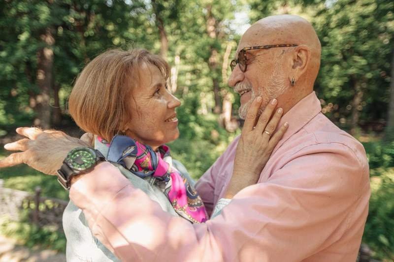 Un couple de seniors souriant s'enlace tendrement dans un parc baigné de lumière, complice et détendu