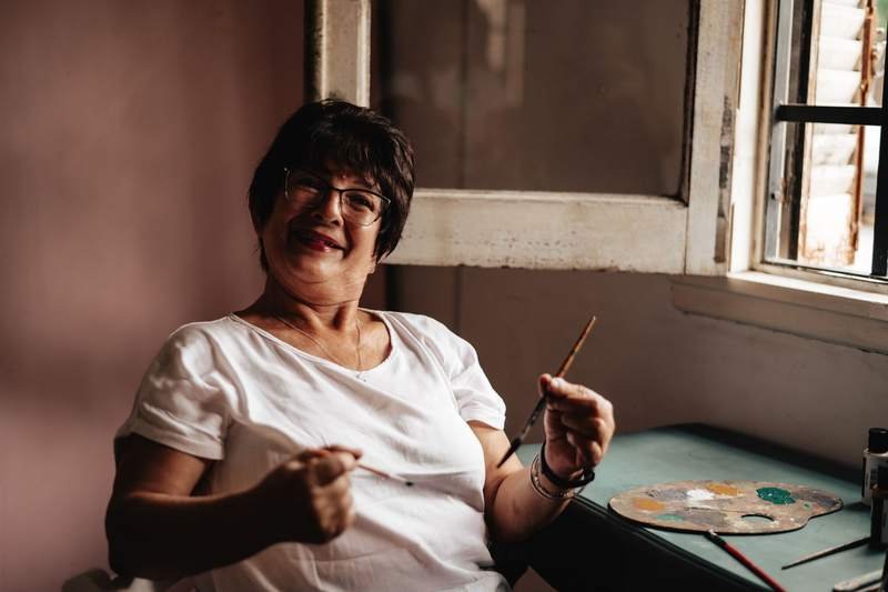 Femme âgée souriante, pinceau à la main, dans son atelier de peinture entourée de toiles et de matériel