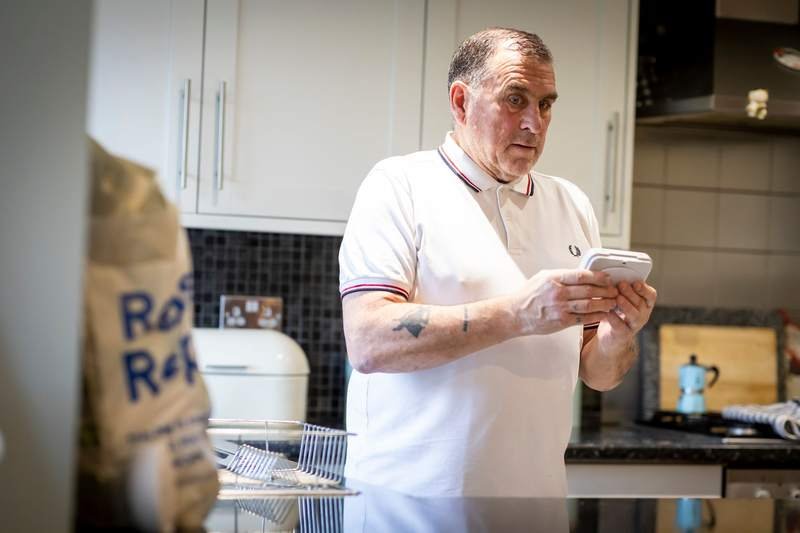 Un homme âgé en polo blanc consulte attentivement son smartphone, debout dans une cuisine moderne et lumineuse