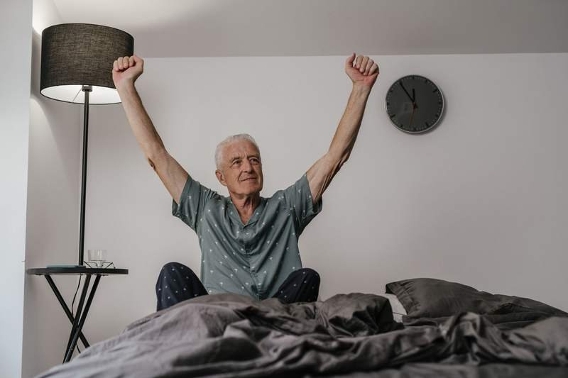 Homme âgé en pyjama assis sur son lit qui s'étire les bras au réveil, dans une chambre claire et paisible