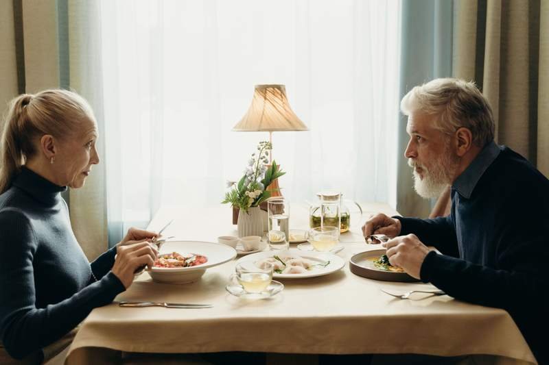 Un couple âgé partage un repas à une table nappée, près d'une fenêtre lumineuse et d'une lampe allumée.