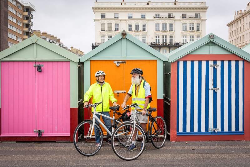 Couple âgé debout avec ses vélos devant une rangée de cabines de plage colorées, en bord de mer, par beau temps