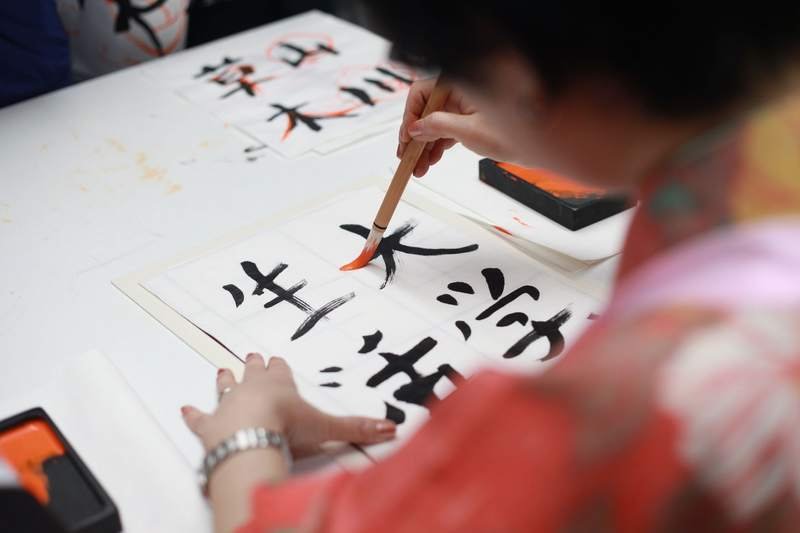 Une main trace des caractères japonais au pinceau et à l'encre noire sur une feuille de papier blanc.