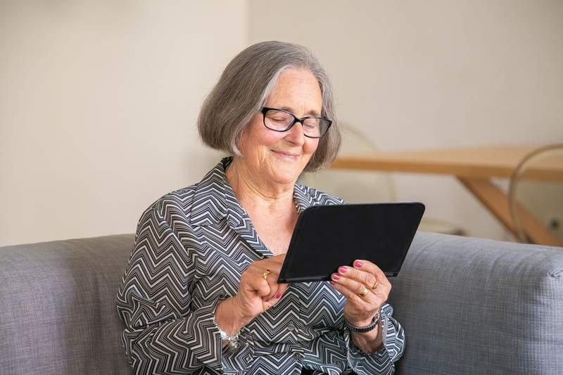 Une femme aux cheveux gris et lunettes sourit en utilisant une tablette, assise confortablement dans son canapé.