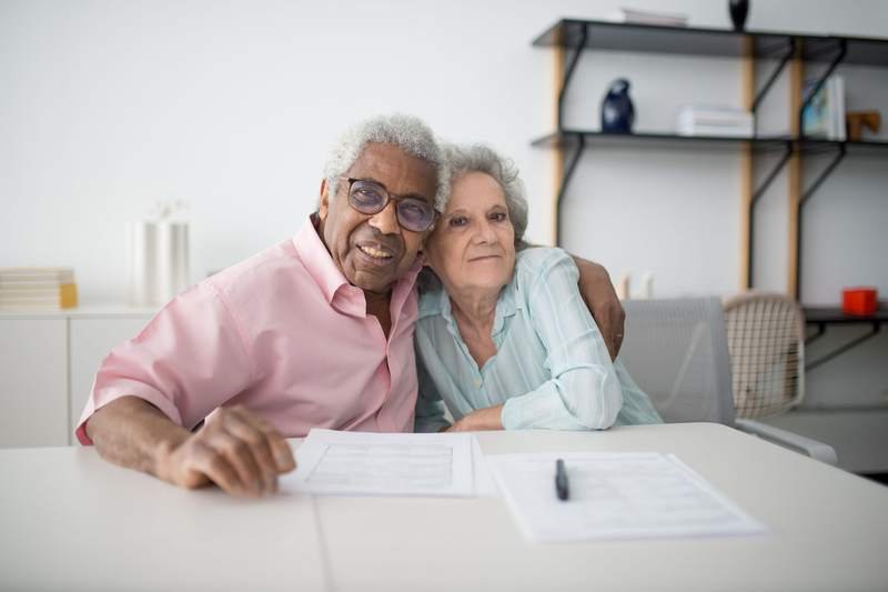 Un couple de retraités, joue contre joue, sourit devant des documents posés sur une table blanche, stylo à la main.
