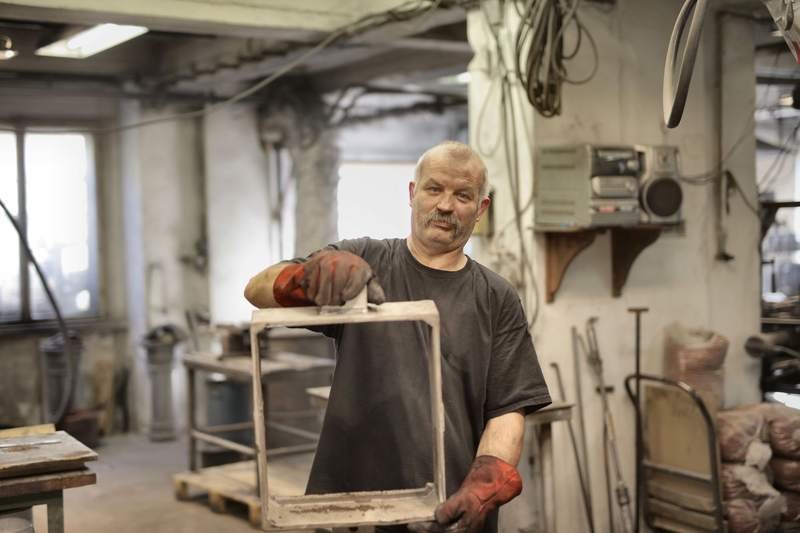Un artisan aux cheveux blancs présente une pièce qu'il vient de façonner dans son atelier encombré d'outils.