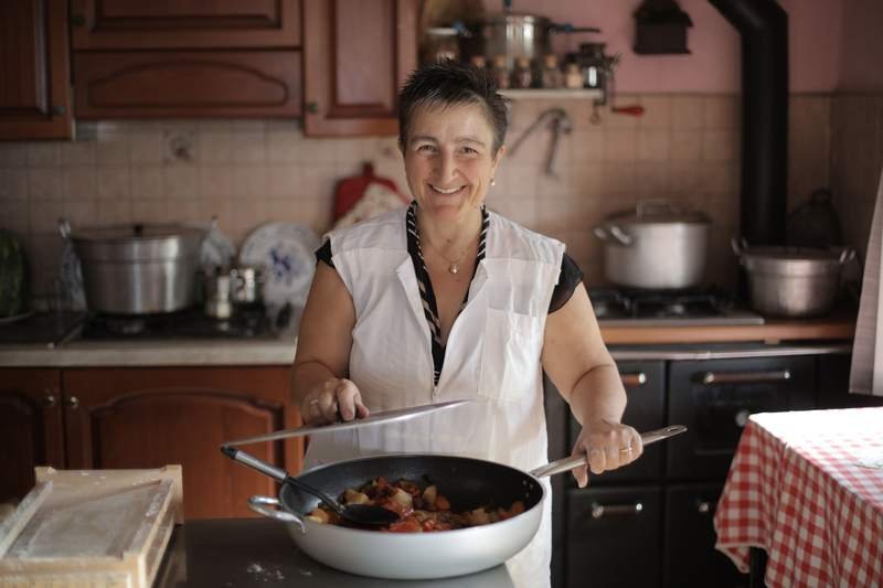 Femme aux cheveux blancs souriante, en chemise blanche, dans sa cuisine chaleureuse avec le repas qu'elle a préparé