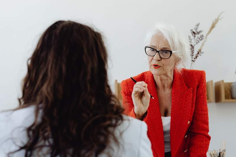 Une femme aux cheveux blancs en blazer rouge échange avec une interlocutrice lors d'un rendez-vous professionnel.