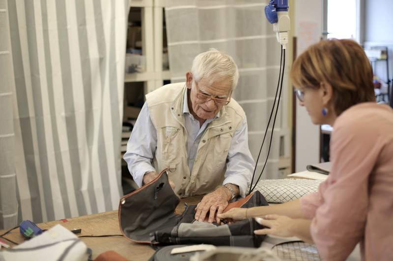 Un artisan âgé travaille le cuir sur son établi pendant qu'une cliente observe, dans un atelier encombré.