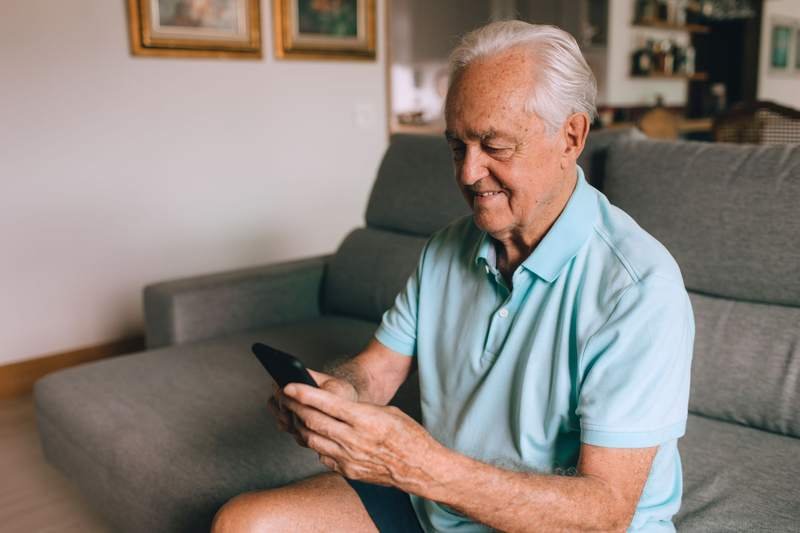 Un homme âgé assis dans son canapé consulte attentivement l'écran de son smartphone.