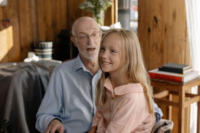 Un grand-père assis dans un fauteuil rit avec sa petite-fille installée sur ses genoux, dans un salon chaleureux.
