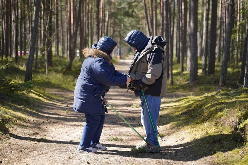 Un couple âgé en manteaux d'hiver règle ses bâtons de marche sur un chemin forestier bordé de pins.