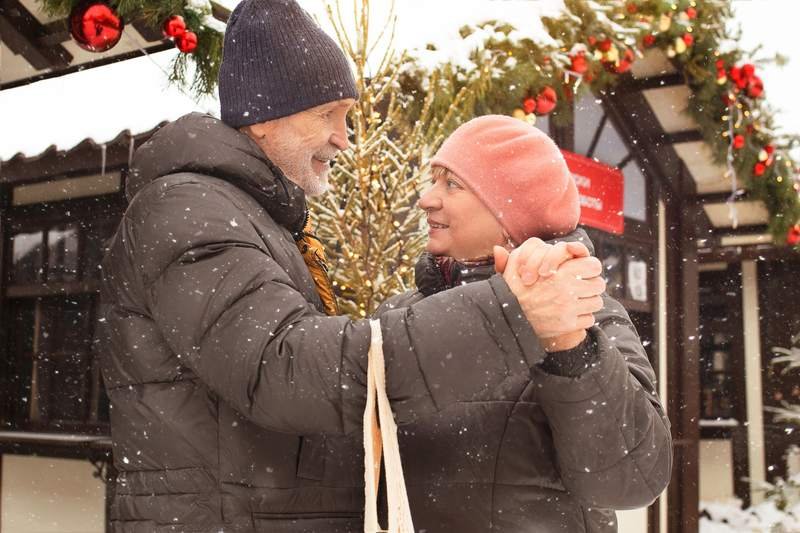 Un couple âgé en doudounes et bonnets se sourit dans la neige, devant un décor de fête hivernal.