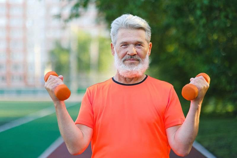 Homme âgé en tenue de sport soulevant des haltères orange en plein air, sur une piste de stade au soleil