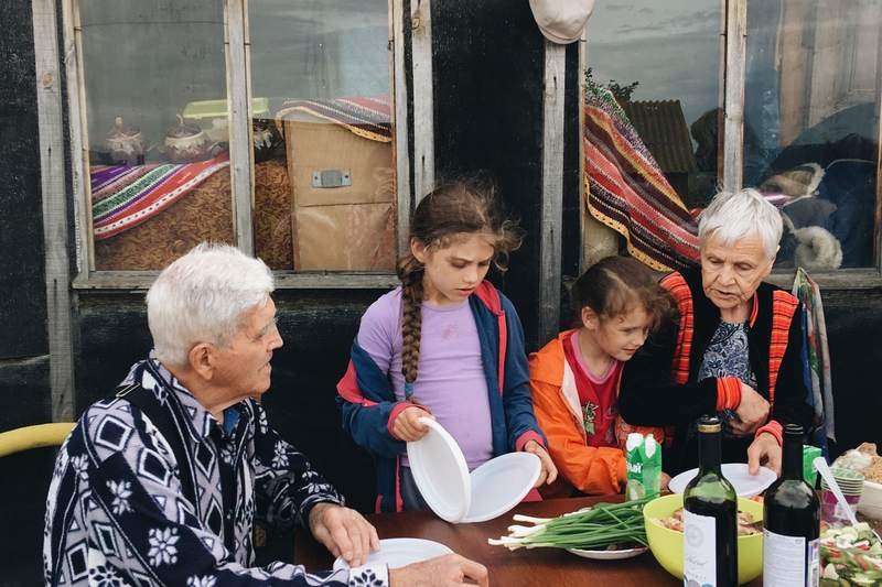 Des grands-parents et leurs petits-enfants dressent ensemble une table en plein air, sous un auvent, pour le repas.