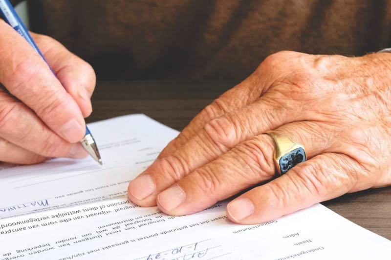 Gros plan sur la main d'une personne âgée portant une alliance en or qui signe un document au stylo