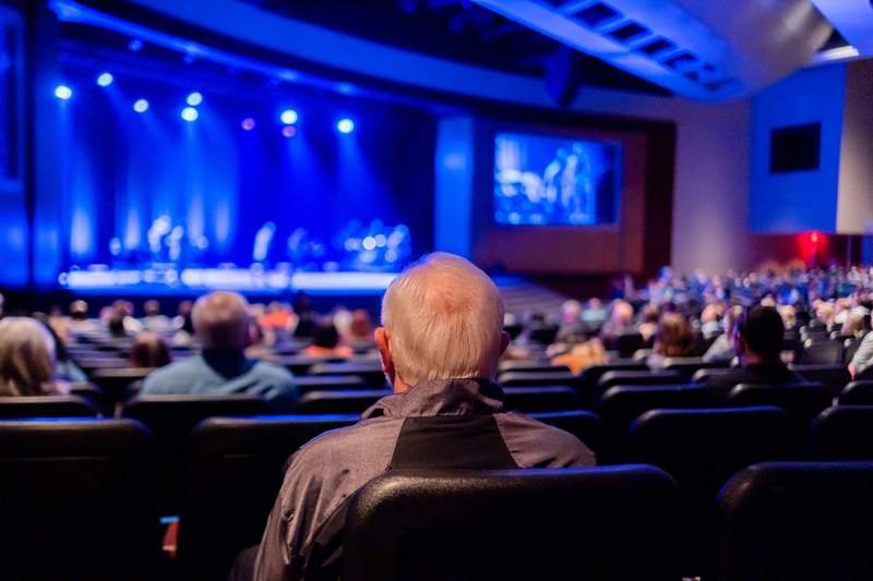 Un spectateur aux cheveux gris, vu de dos, assiste à un spectacle dans une salle pleine éclairée de bleu.