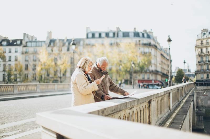 Un couple âgé consulte un plan au bord d'un quai, dans une ville européenne aux immeubles haussmanniens.