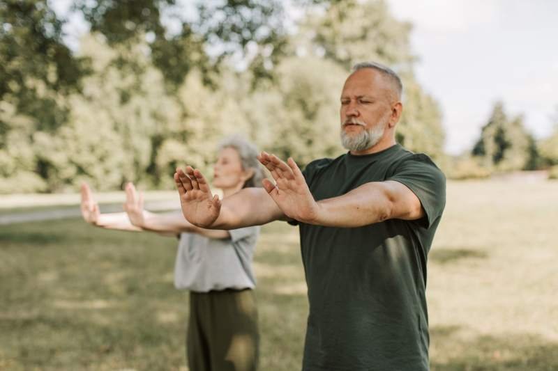 Une femme et un homme âgés pratiquent un mouvement de tai-chi, bras tendus, dans un parc ensoleillé.